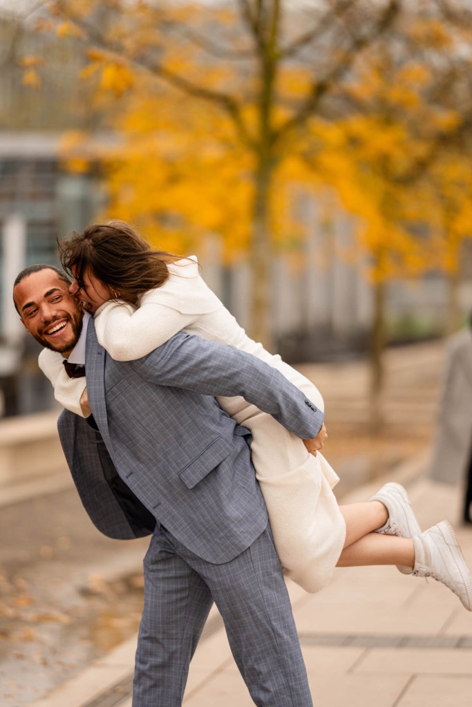 Authentische Paarfotografie Herbst Köln – Mann trägt Frau lachend durch die Straßen © Chirlyart